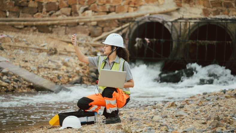 Field worker assessing water flow near a concrete drainage outlet